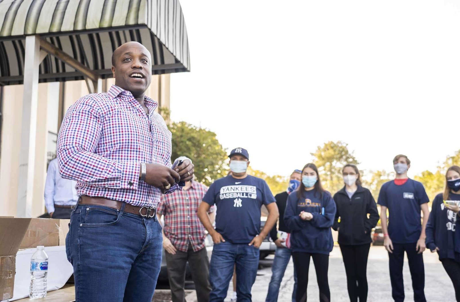 A bald man in jeans and a purple button down shirt stands in front of a group of masked people