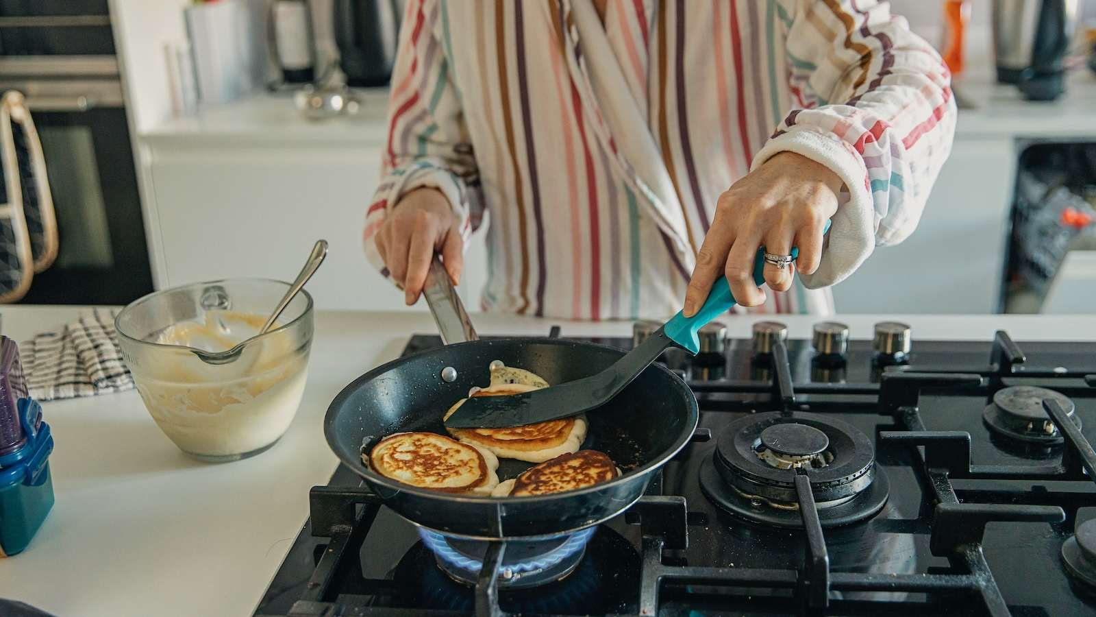 A woman is shown making pancakes in a skillet on a natural gas-burning stove.