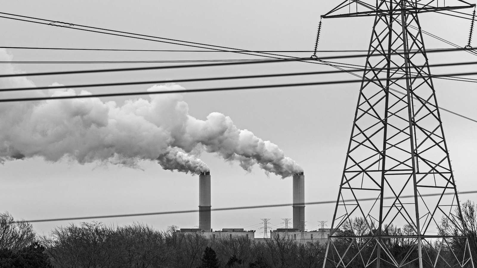 Black and white photo of smokestacks in the distance, with transmission lines in the foreground