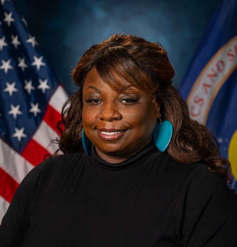 A woman poses in front of a blue background with the U.S. flag (left) and NASA flag (right) behind her.