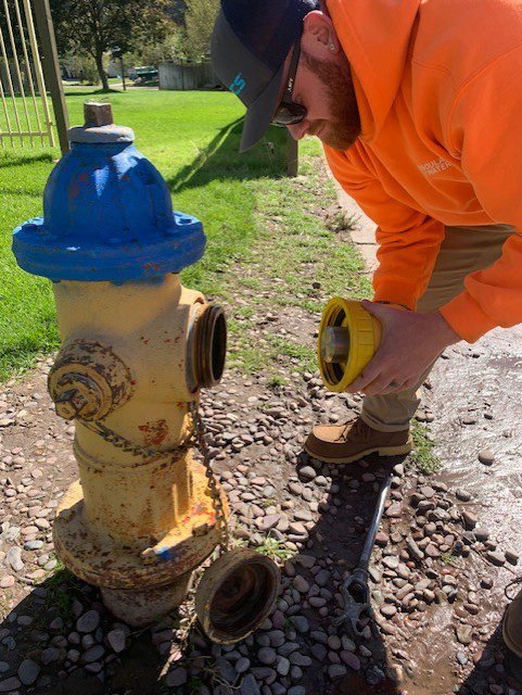 A man crouching next to a fire hydrant, holding some kind of cap attachment that has been removed from the hydrant and inspecting, possibly, the whole assembly