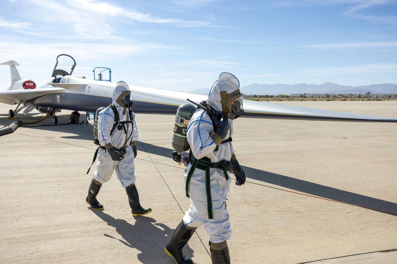 Two maintainers in white protective suits, air tanks, masks, rubber gloves, and boots walk around the front of NASA’s X-59 quiet supersonic research aircraft during a hydrazine safety check at U.S. Air Force Plant 42 in Palmdale, California, on Aug. 18, 2025. The sky is mostly clear, with some vegetation and mountains visible in the background. The aircraft has its canopy open, a ladder positioned on the right side of its fuselage, and air ducts extending from the aircraft. 