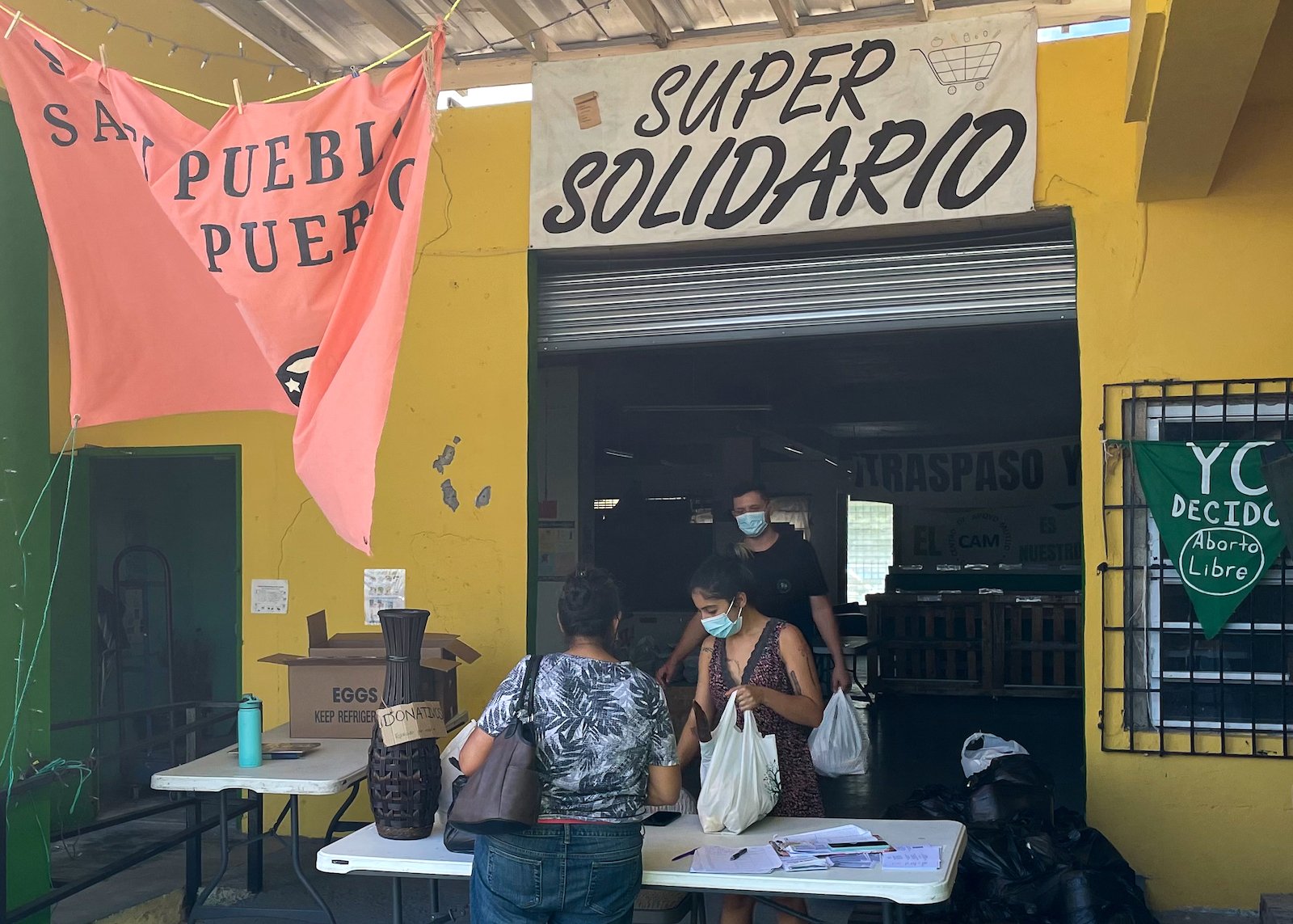 A sign that says 'super solidario hangs above an archway where people are meeting at a table. There is also a red flag with 'solo el pueblo salva al pueblo' on it hanging above them