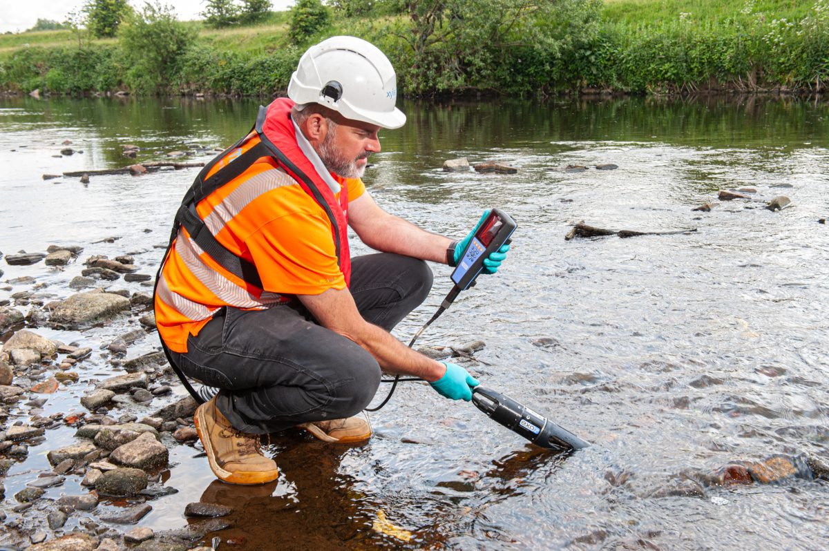 Water industry operative in hard hat and luminous vest squats on bed of river or stream with handheld apparatus, one end of which is a cigar-shaped object that he is dipping into the water, while the other end is a handheld unit that he appears to be looking at, as if inspecting the readout