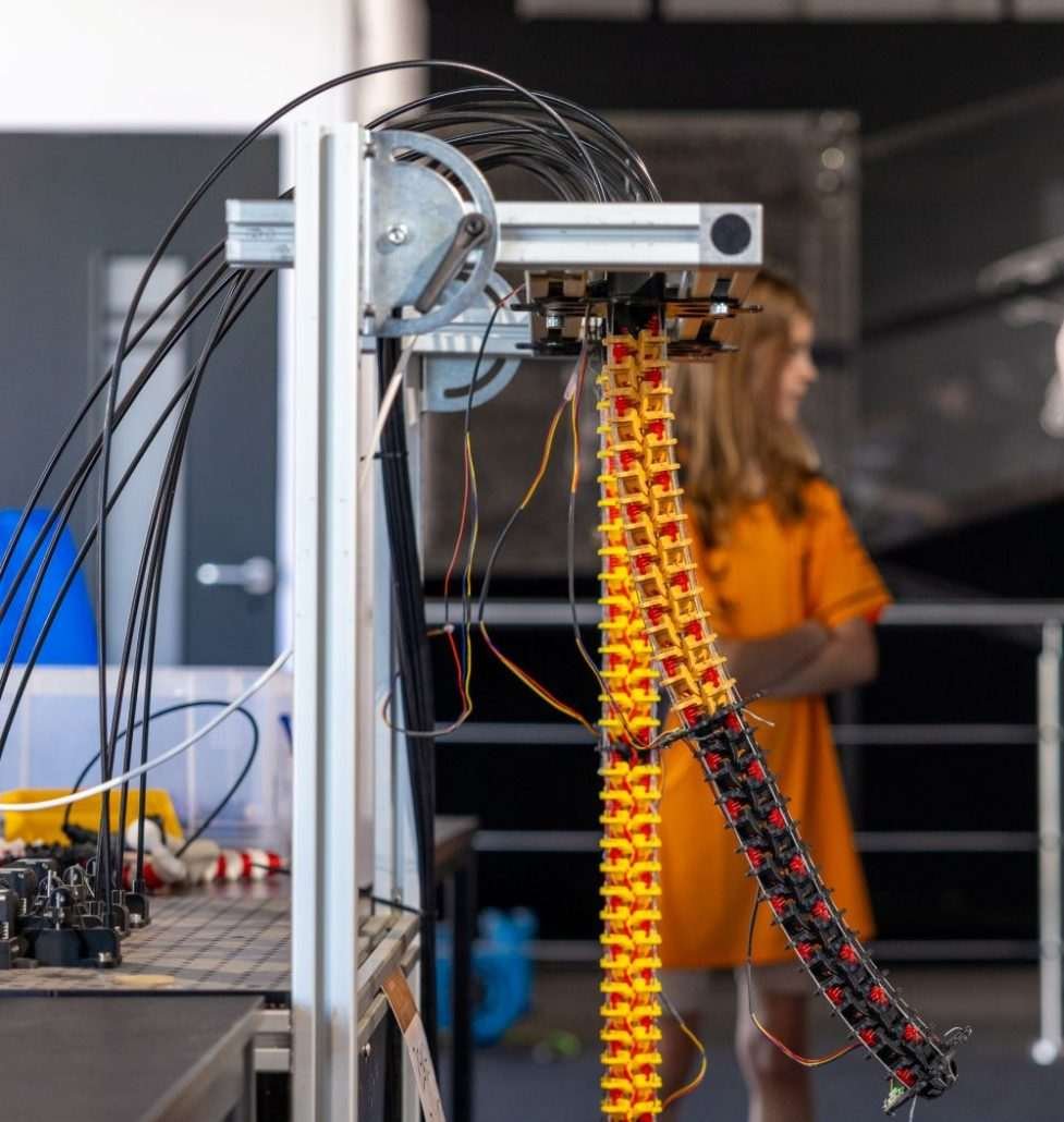 rope-like appendage hangs from upright scientific laboratory mounting equipment with wires and other components visible nearby, while a woman is visible in the middle distance wearing a pink garment