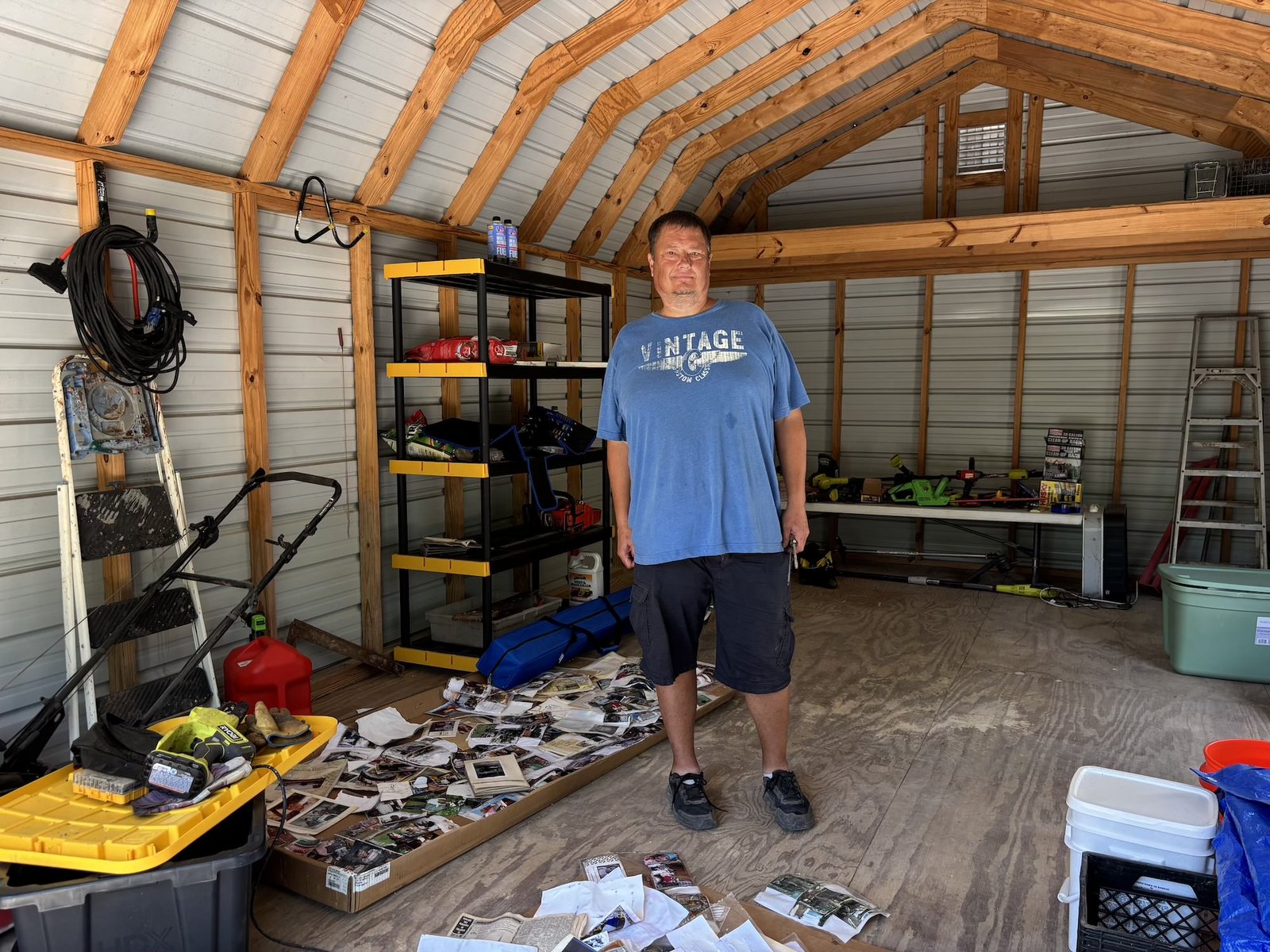 A man in a blue shirt stands in a warehouse or garage with a pile of papers