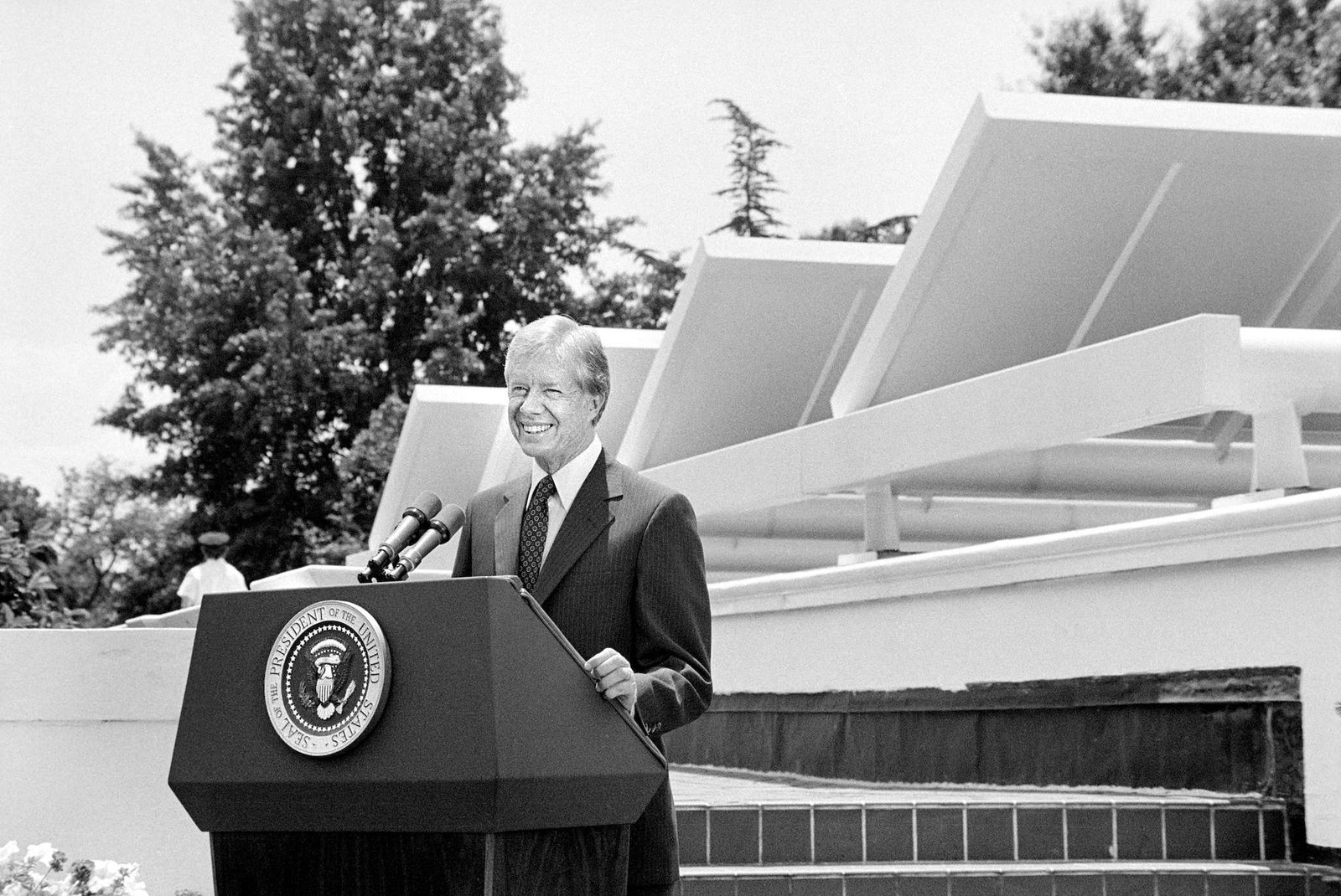 a black and white photo of Jimmy Carter giving a speech behind a podium and in front of several solar thermal panels