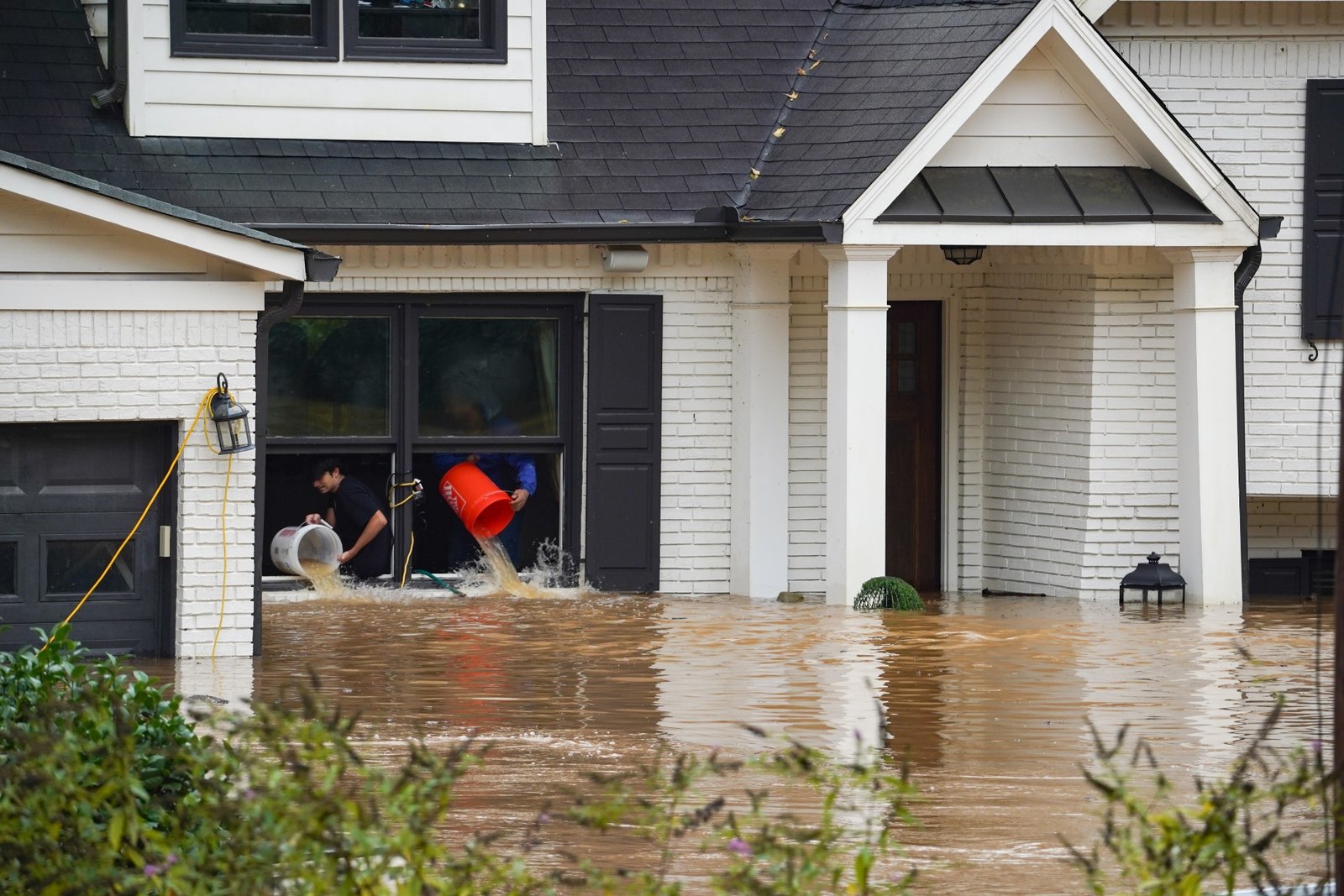 A white brick house is surrounded by muddy water that reaches up to the first floor windows. Two people use buckets to pour water out of the window.