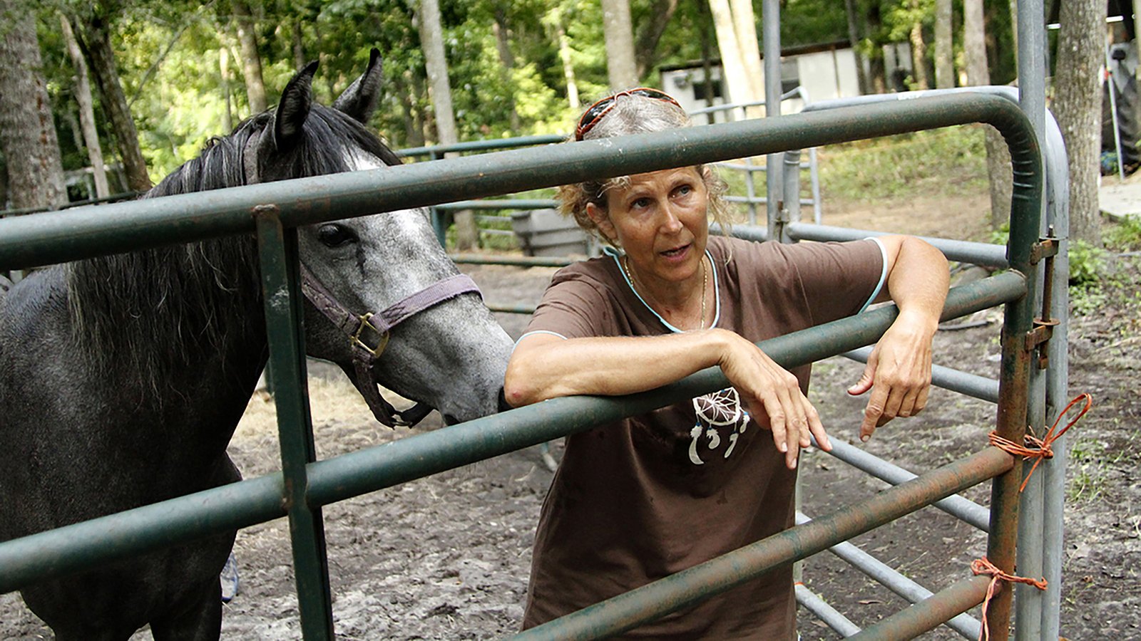 Jane Blais leans against the railing of a horse enclosure as a black horse stands beside her.