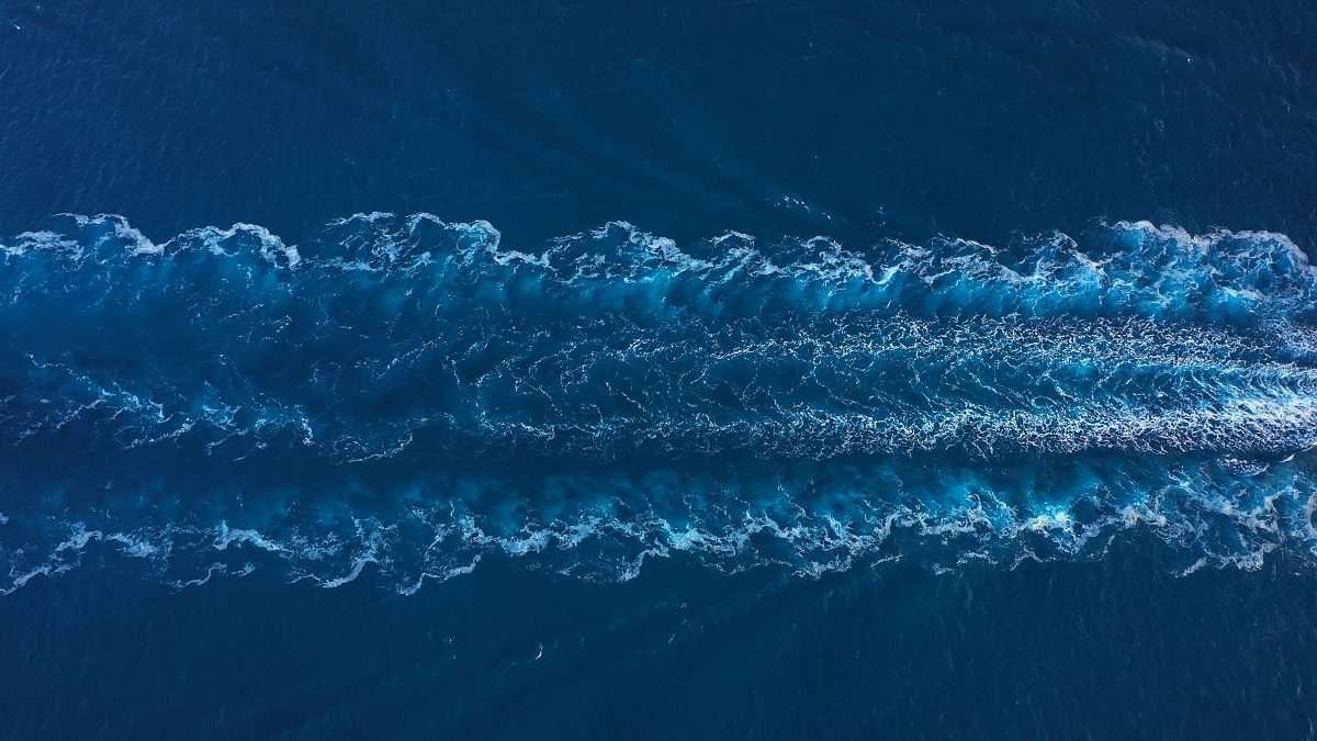 A section of open sea, seen from above, with the image bisected horizontally by the white and pale blue eddies of the wake of a ship, now out of view to the right of the image