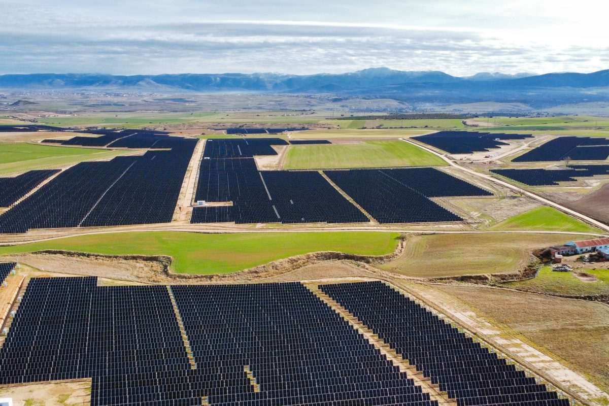View from above of arid unpopulated landscape, resembling a desert region, with dark mountains in the distance, and large geometric segments of the land appearing appearing as a darker colour because populated with solar panels