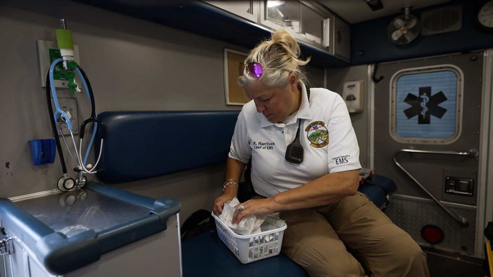 A woman in a white shirt and walkie talkie sits in an ambulance rummaging through a box