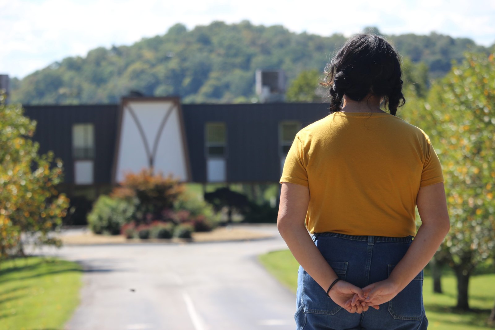 A woman in a yellow tee shirt and jeans with her hair in pigtails stares at a building in the distance