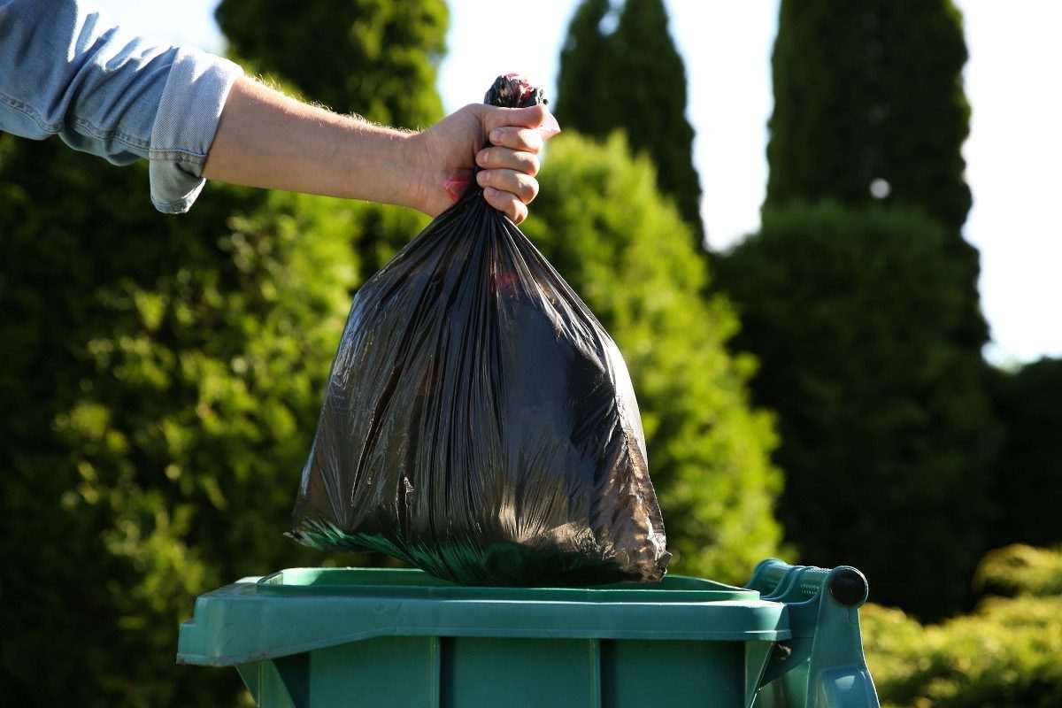 A human arm puts a bulging black bag into a green wheelie bin, in an outdoor location with trees in the background