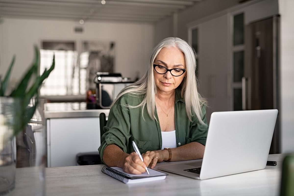 A woman in smart-casual attire sitting behind a desktop with a laptop on its surface in tasteful, minimalist home or office or kitchen-like space, with houseplant just visible at the edge of the image, and subdued lighting as though daytime in a temperate geographic region