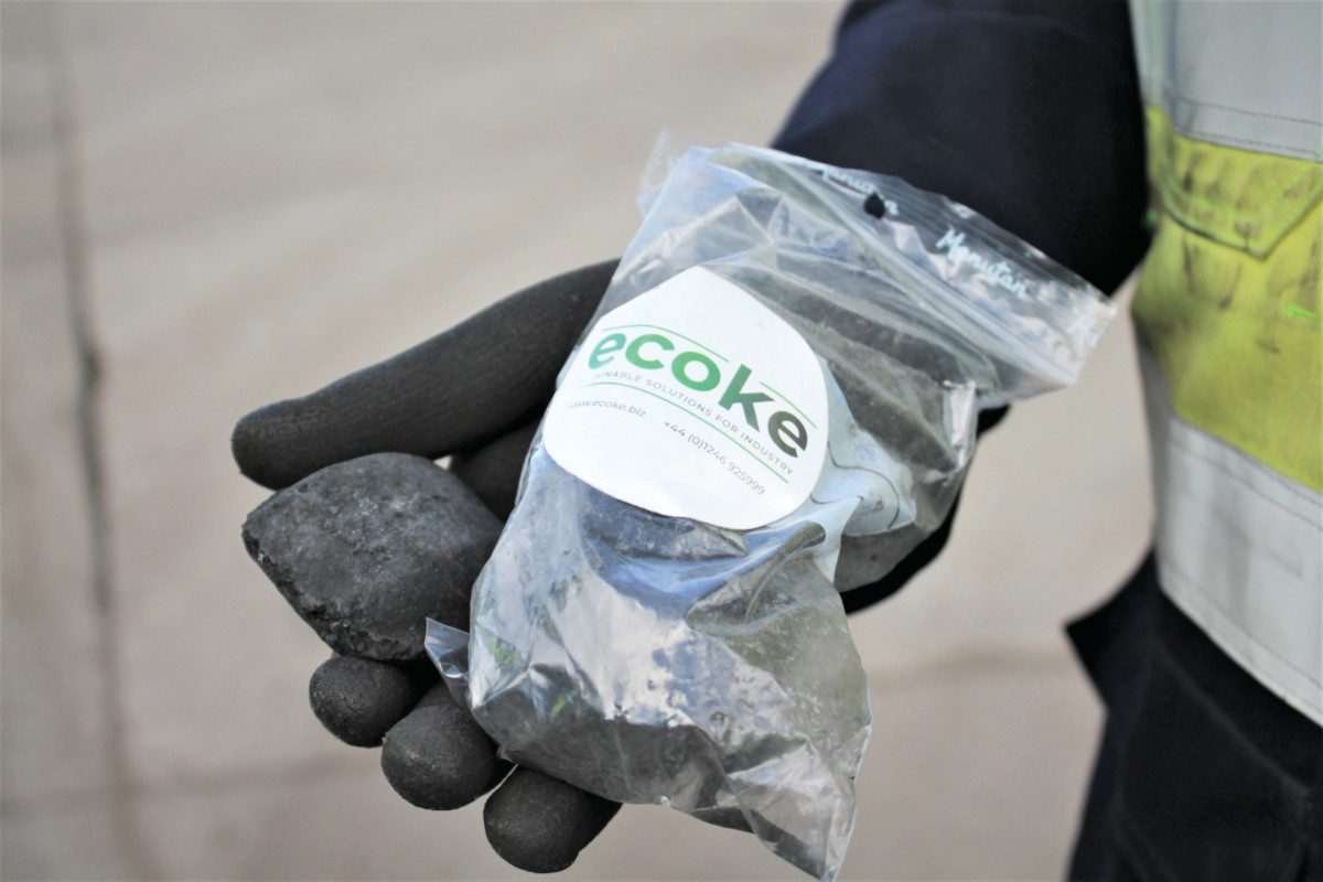 A gloved hand holds a transparent bag bearing the label 'ecoke' and containing black, coal-like briquettes