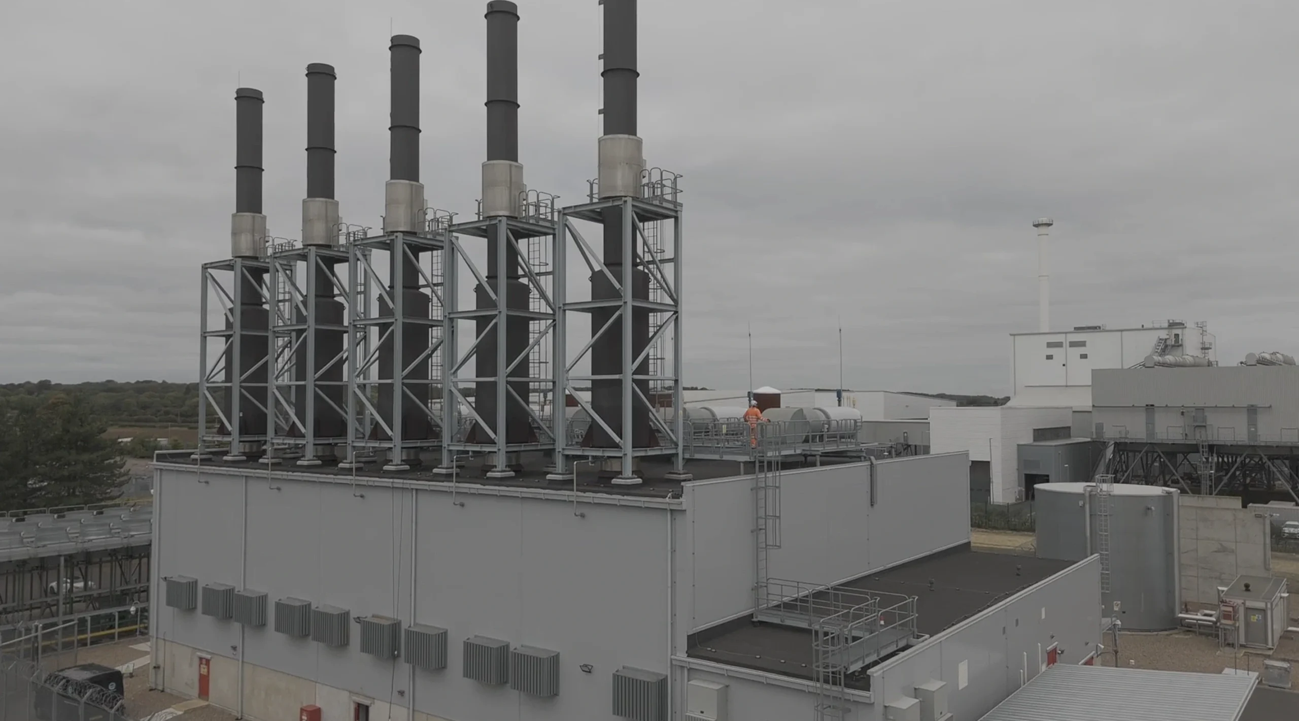 View from slightly to the side and above the rooftops of a large cuboidal building upon the roof of which are arrayed five chimney-like structures with nearby multi-tiered personnel platforms, all within an outdoor landscape of little note, with heavy cloud cover and dark-coloured ground in the background, so that the whole image appears somewhat grey or dull in tone