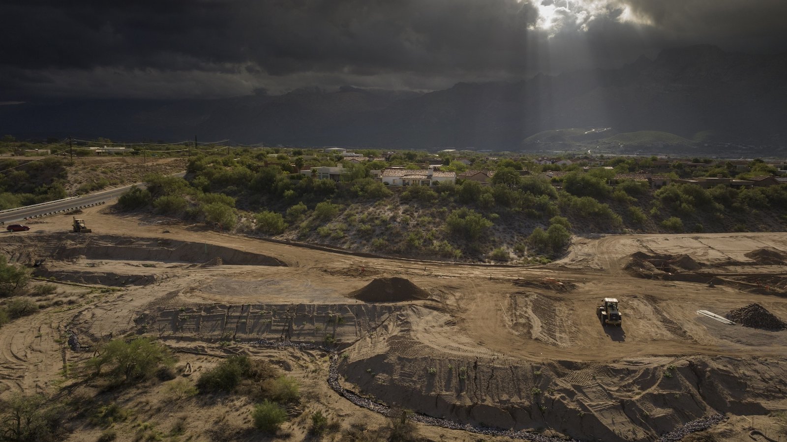 a construction site next to houses in the southwest. Above, clouds create moody lighting with a slight sunbreak over a piece of construction equipment