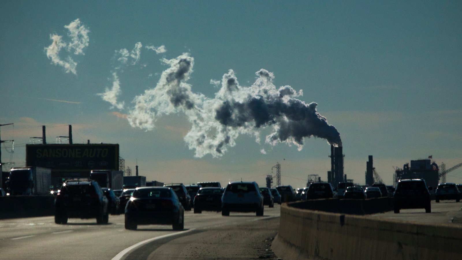 Cars drive on a highway towards an industrial complex belching smoke