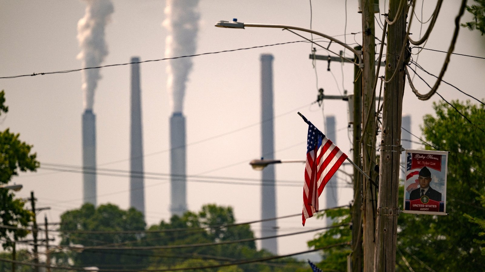 Several smokestacks stand in the background with one belching smoke and in the foreground are trees and an American flag