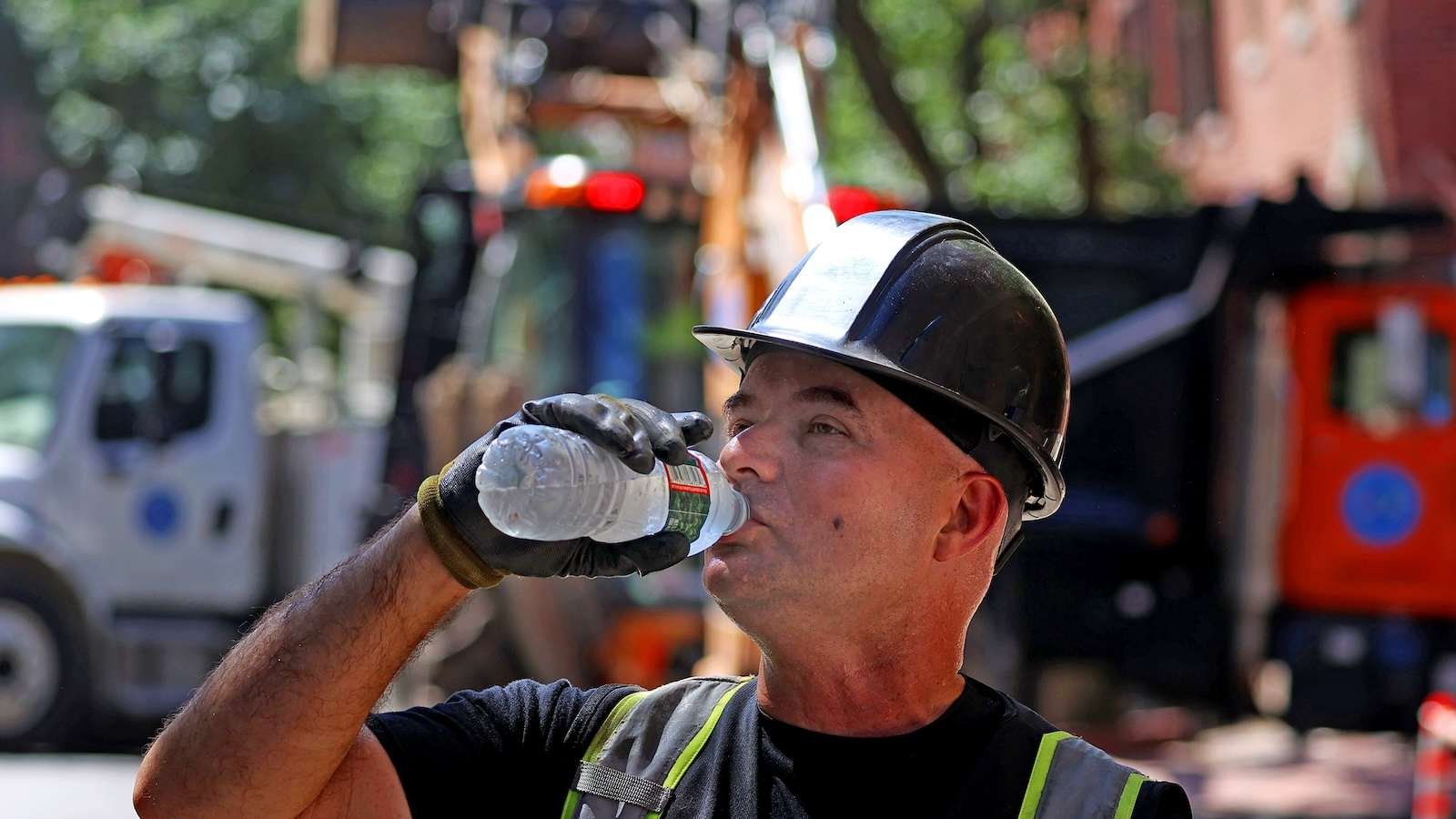 a construction worker with a hard hat on drinks from a cold bottle of water in front of street construction