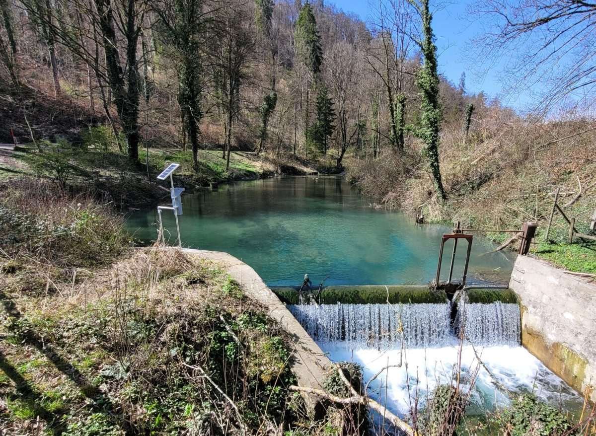 A rod-like, upstanding structure with a visible solar panel at its summit, sits upon a rock in close proximity to a river weir, with water visibly falling from a short height, while in the background is visible sparse, brown, dessicated woodland