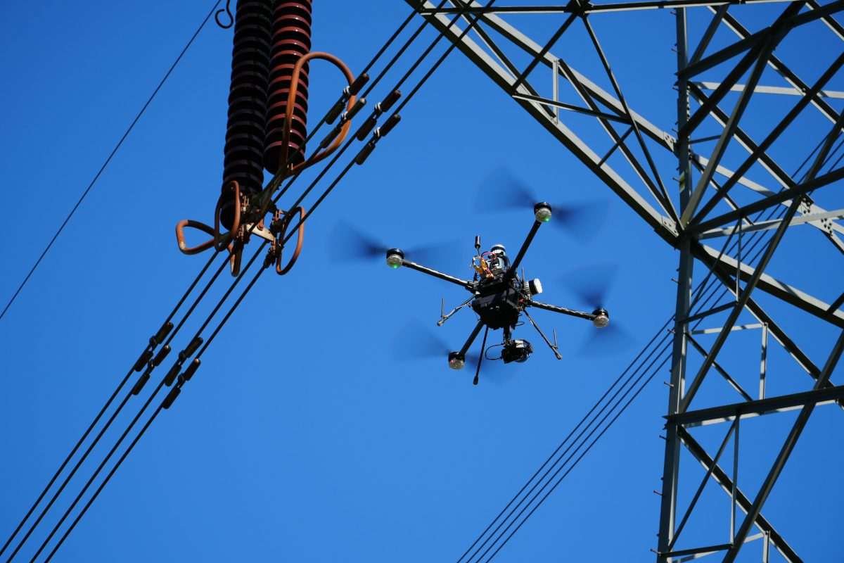 A drone in mid-air near an electricity pylon, with a small portion of this structure in view, all against a blue sky background