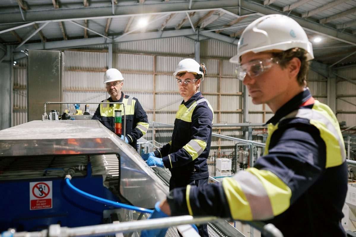 Workers in hard hats and protective eyewear stand atop a walkway area in an industrial facility looking at cuboidal equipment assembly on a benchtop in the foreground