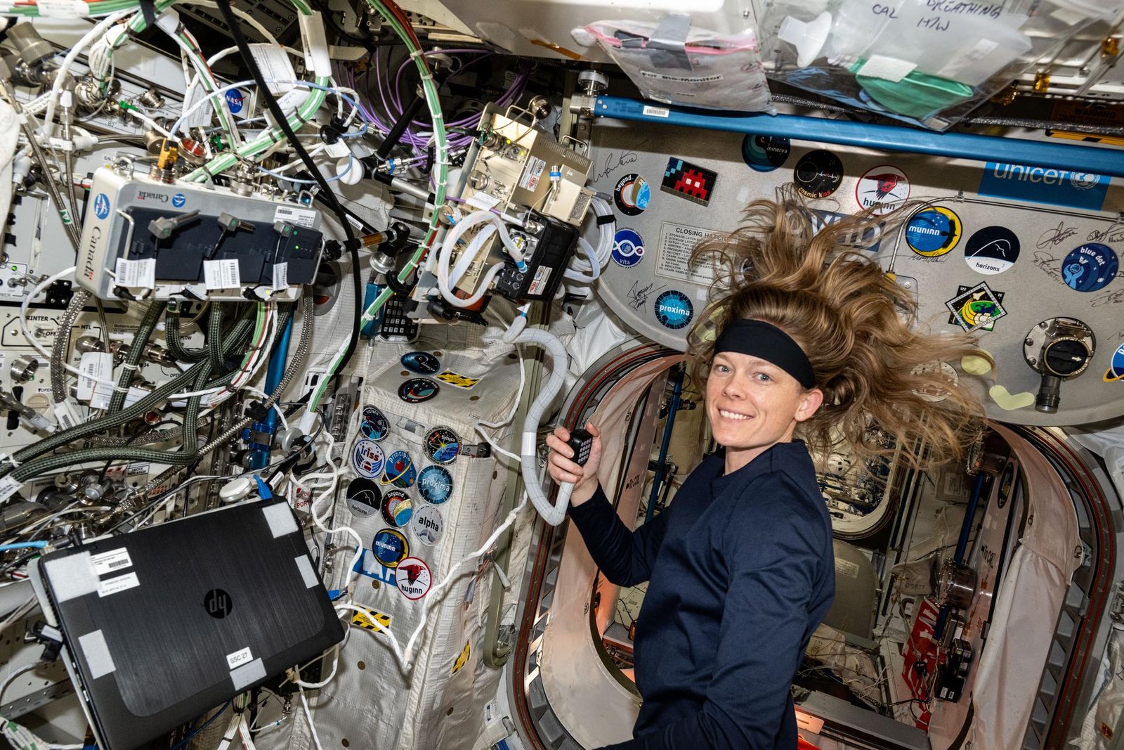 Nichole Ayers smiles at the camera while holding a hand-held radio aboard the space station. Her long blonde hair floats upward in microgravity. To her left, a laptop and cables are mounted on the wall, and the wall behind her is decorated with several mission stickers.