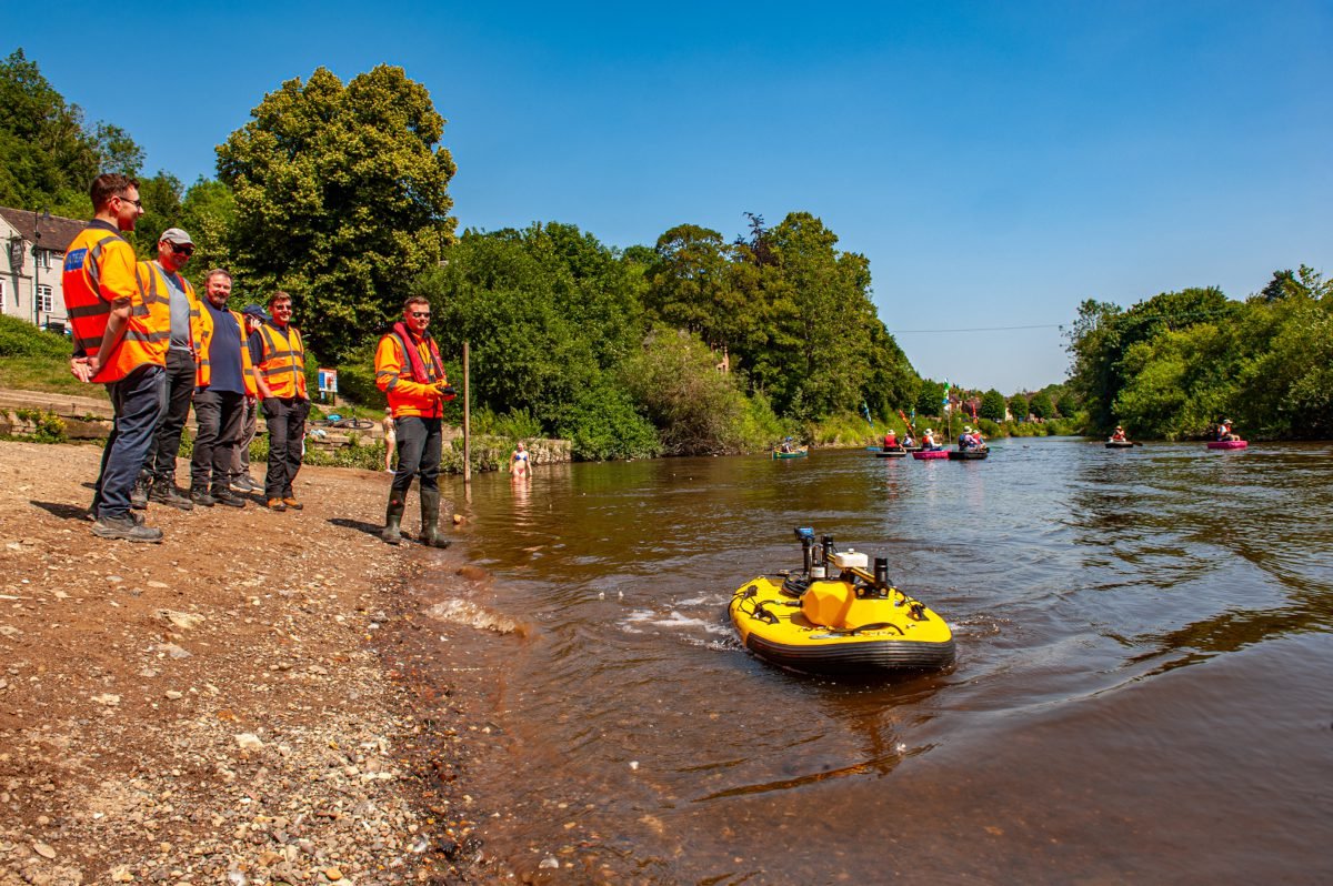 Several people in luminous work vests stand on river bank observing unmanned surface vehicle floating in the shallow region where the river meets the shore, with surroundings also revealing canoists on the river in the distance, surrounding woodland, and part of a house or building