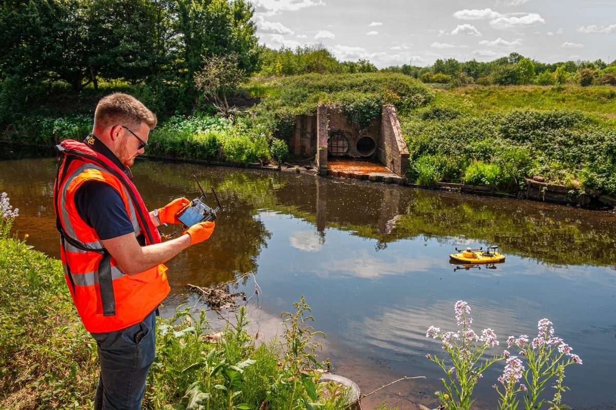 Man in luminous work vest operating a remote control while standing on a river bank, with unmanned surface vehicle visible near the central lane of the river, with background also revealing opposite river bank and surrounding rural land and the sky 