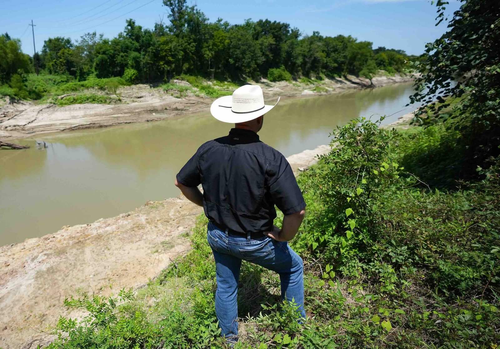 A man in a cowboy hat stands on the bank of a river, with his back to the camera
