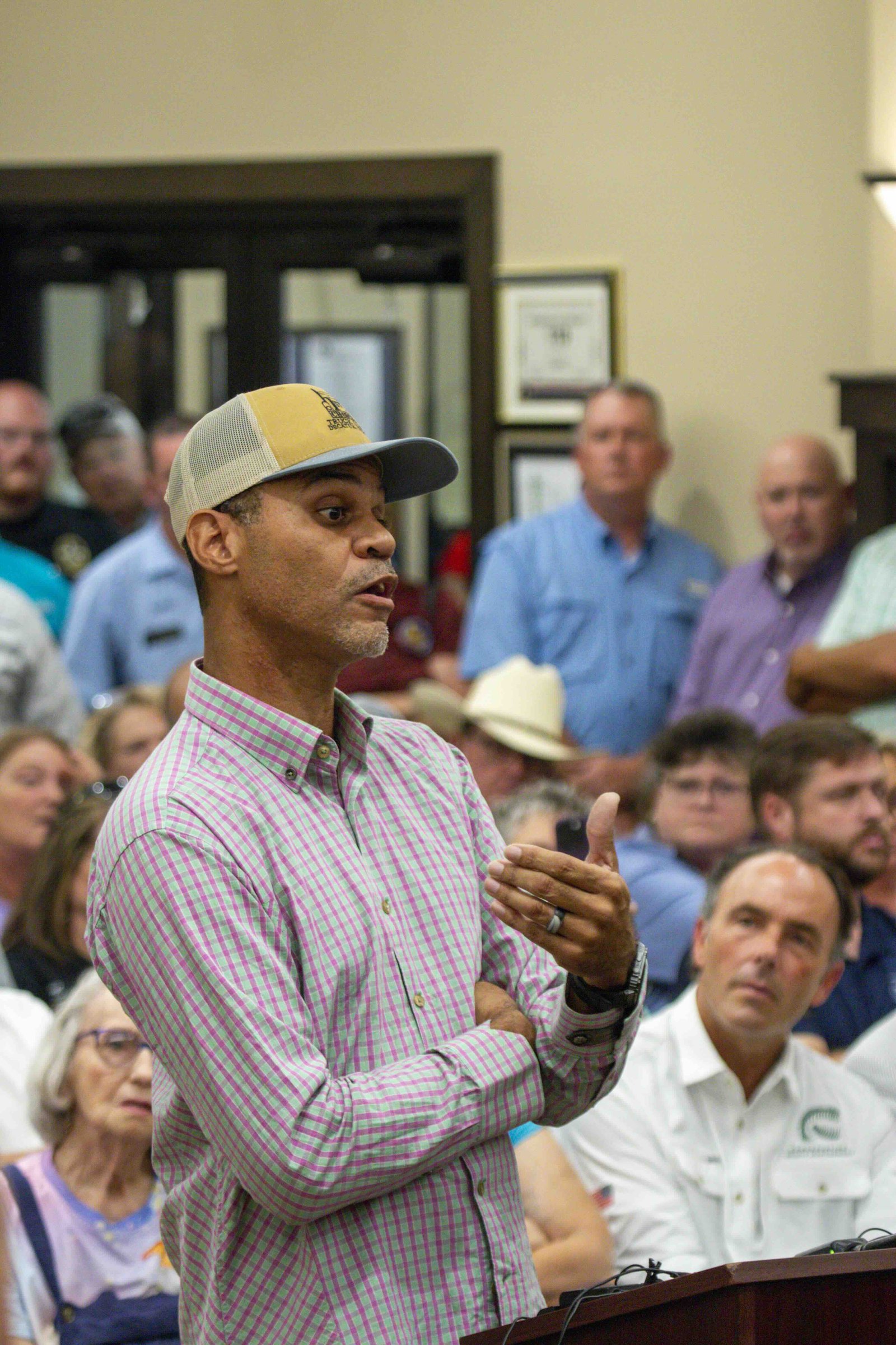 A photo shows a man in a ball cap speaking at a podium in front of a crowded room