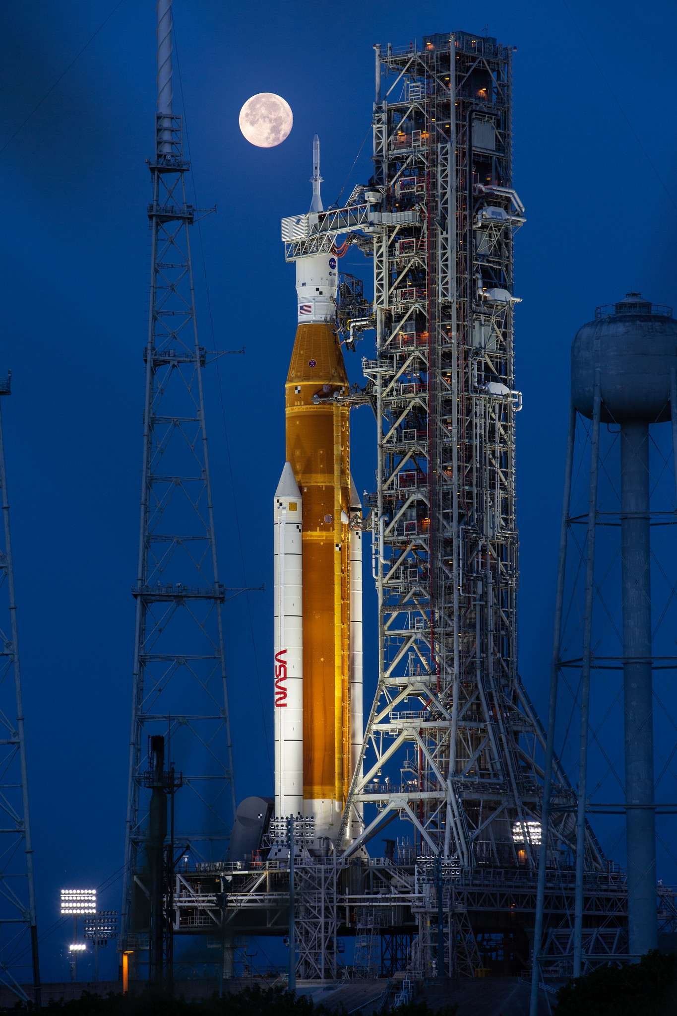 A large orange NASA rocket with white booster rockets stands vertically on a launch pad, lit by spotlights, with the full Moon and night sky behind it.