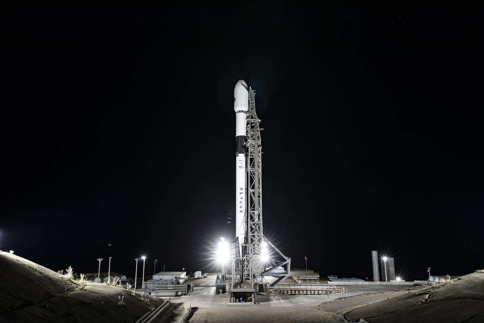A wide-angle, distant photo of a white rocket on the launch pad, brightly lit, with a black night sky behind it.