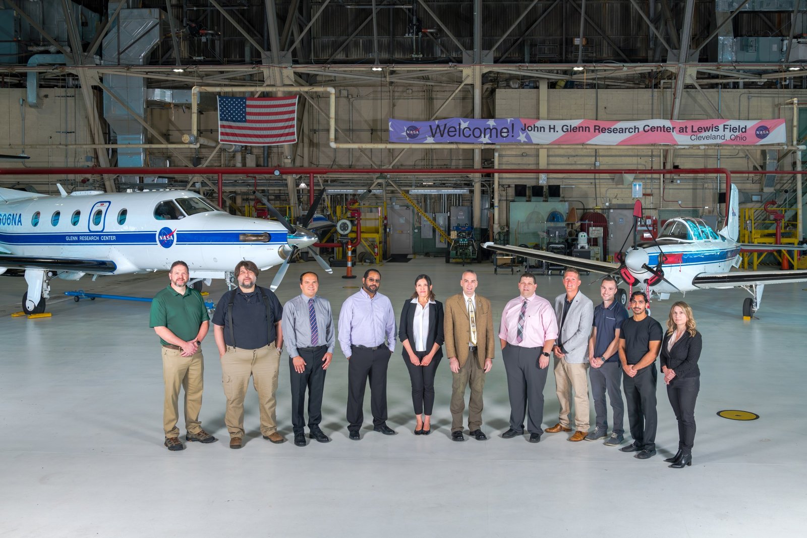 Eleven people stand next to one another inside an airplane hangar. Two small planes sit behind them. An American flag and a banner reading “Welcome! John H. Glenn Research Center at Lewis Field, Cleveland, Ohio” are visible behind the group.