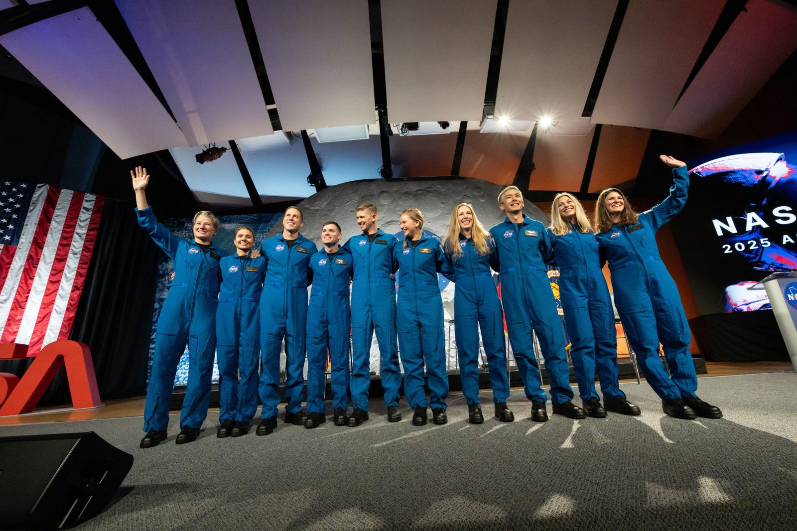 Ten people in blue flight suits wave to an audience in an auditorium.