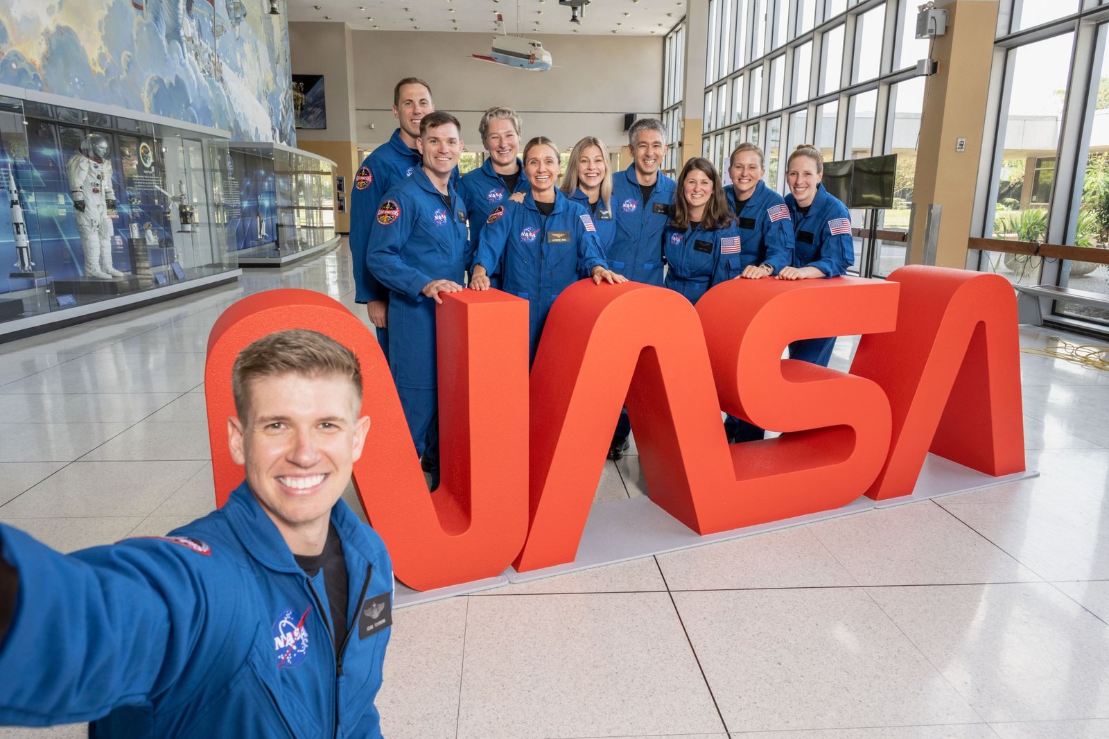 A man in a blue flight suit takes a selfie with a group and the NASA worm insignia in the background.