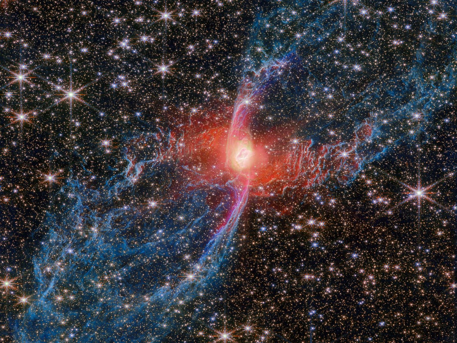 A multicolored nebula stands out against the star-studded background of space. The nebula’s central star is hidden by a blotchy pinkish cloud of dust. A strong red light radiates from this area, lighting up the nearby dust. An elongated purple ‘S’ shape is centered on the heart of the nebula, while two blue lobes stretch diagonally outward to the edges of the image, making the entire structure look like a tilted hourglass.