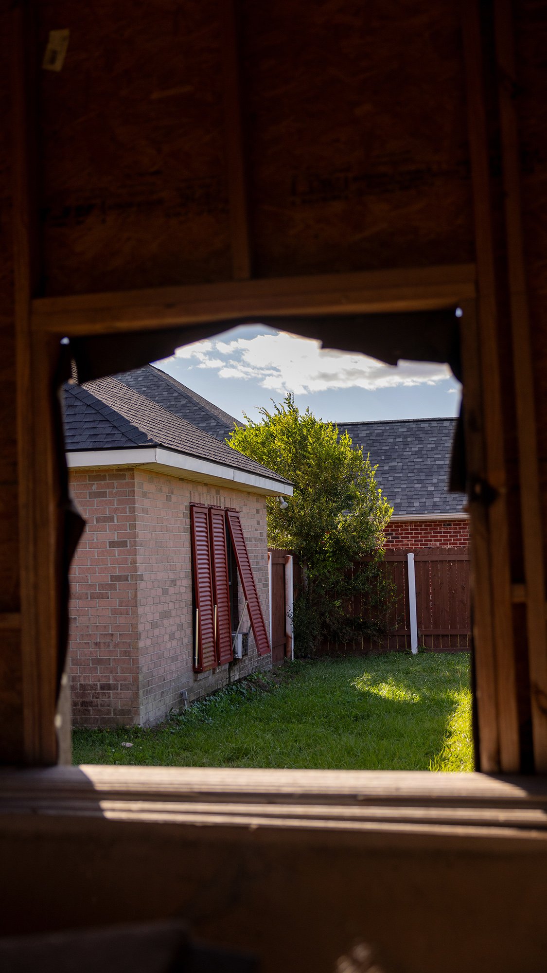 A view out the window of a garage with damaged glass hung instead with mesh. Through the frame, a brick house with loose shutters can be seen