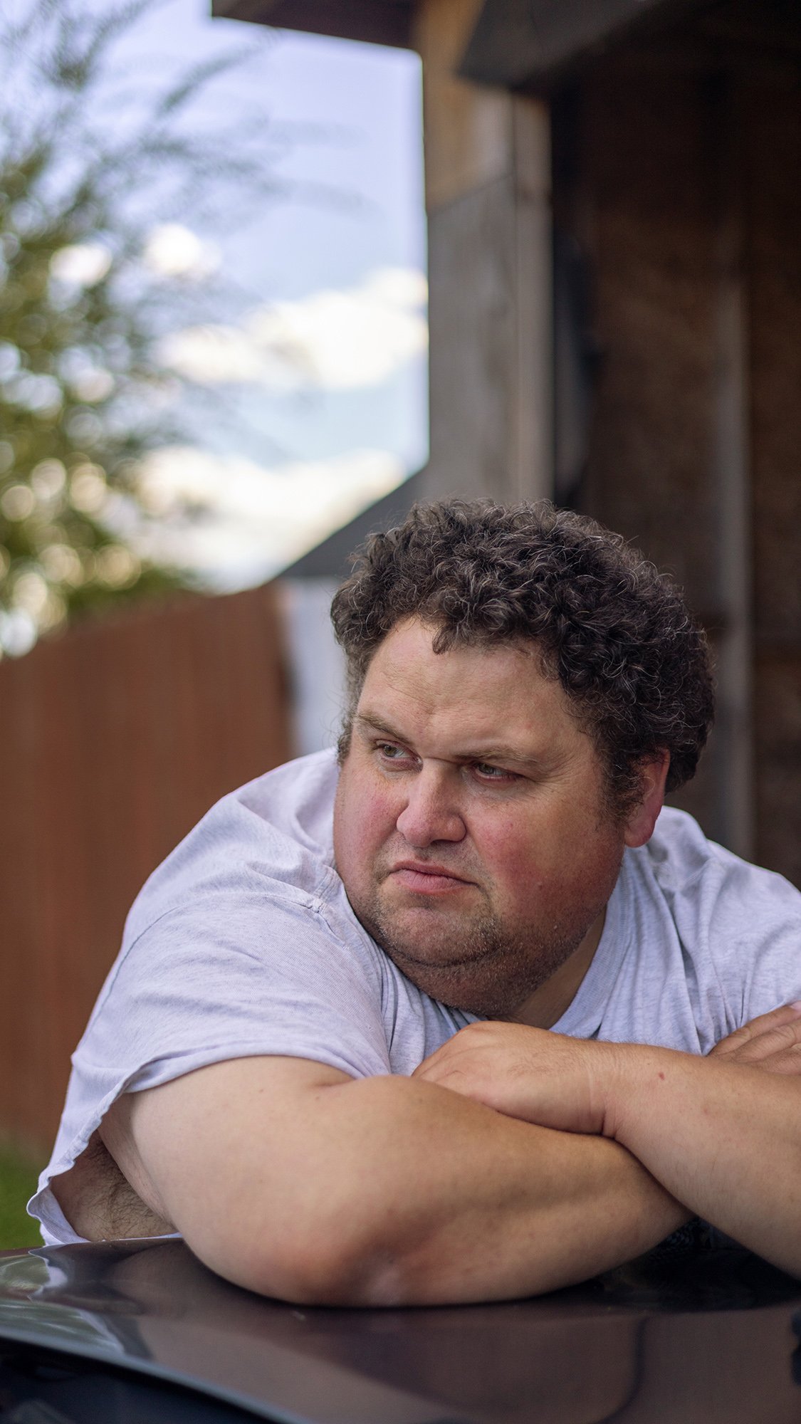 A middle-aged man crosses his arms on a car hood near a storm-damaged house