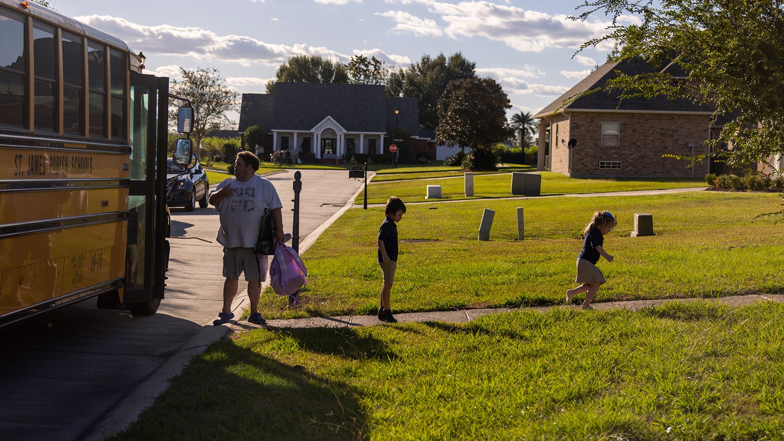A middle-aged man stands near a parked yellow school bus while two children run away from the bus up the walkway of a home surrounded by green lawns