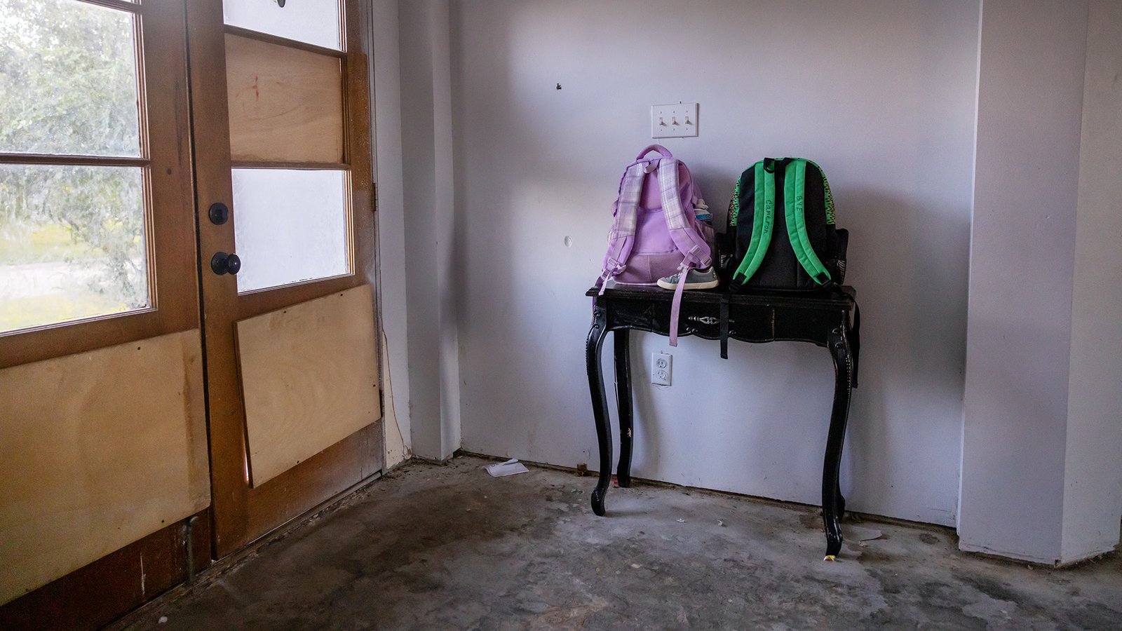 two child-sized backpacks in bright colors stand on a hallway entry table in a home with damaged floors and walls