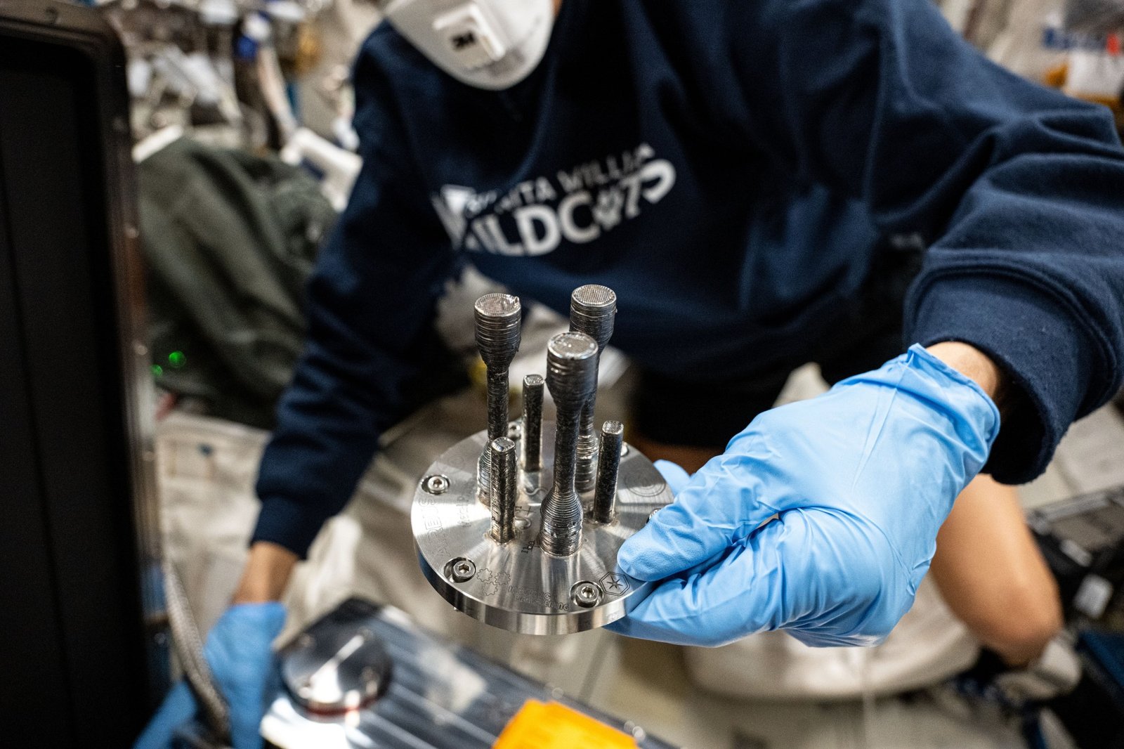 A space station crew member holds the first metal part that was 3D printed in space.