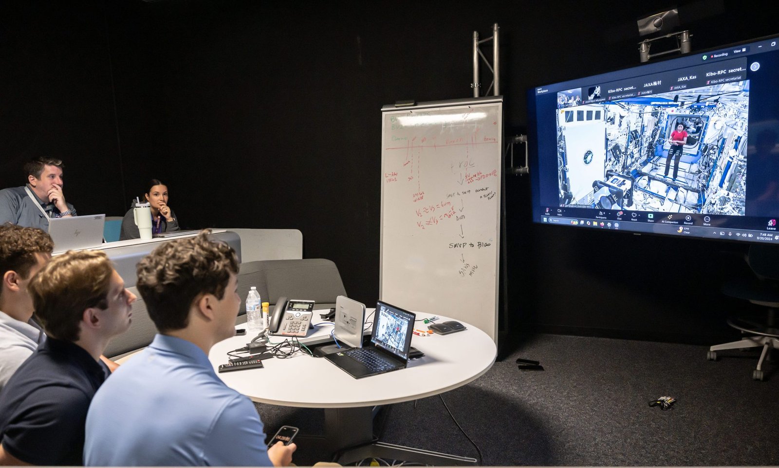 A group of people sit around a circular table in a dark room, engaged in a video call displayed on a large screen showing the interior of a space module with an astronaut visible.