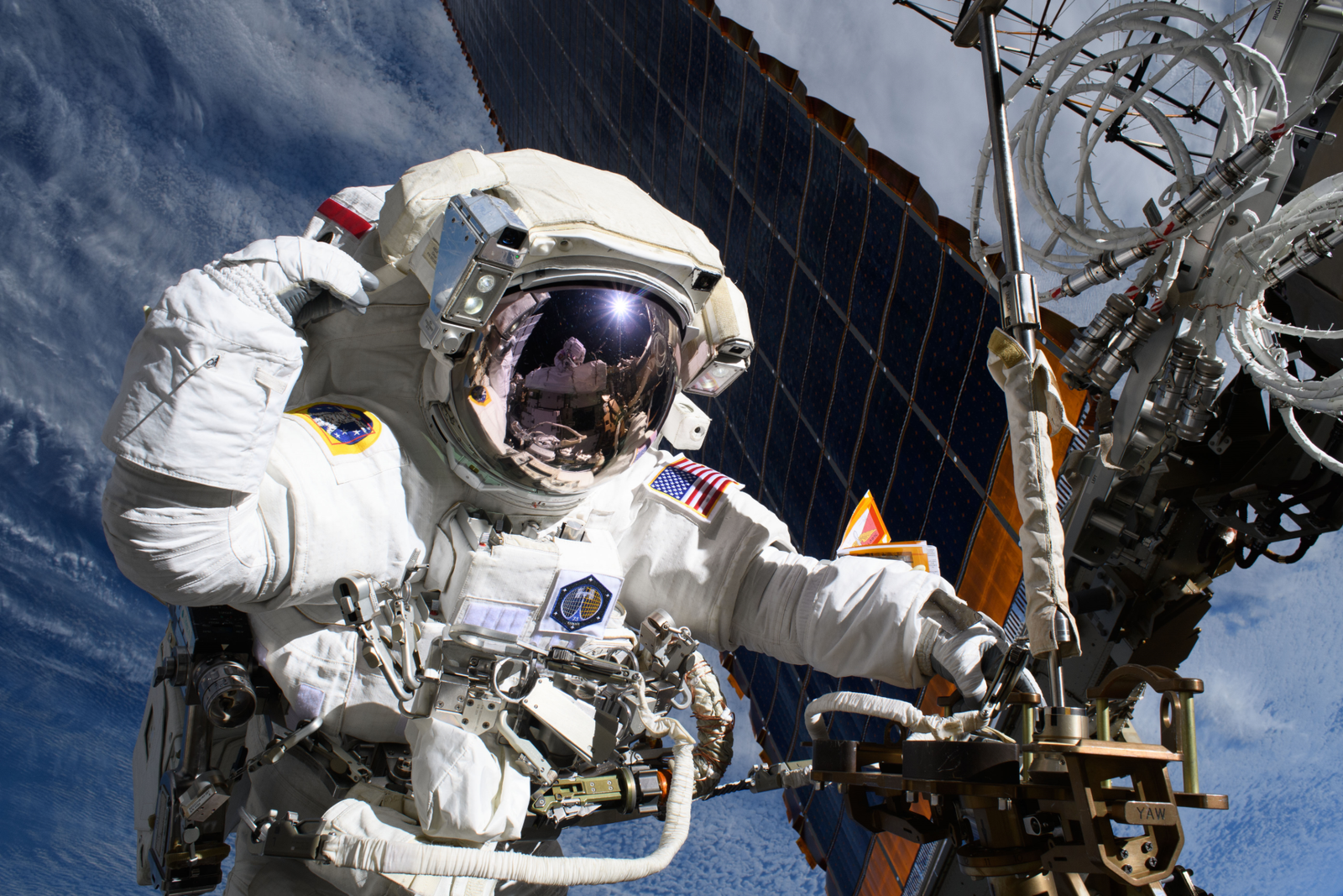 An astronaut outside of the International Space Station has one hand on a truss near a solar panel. Her other hand is by her head. Reflected in her helmet is astronaut Nichole Ayers, also in a white spacesuit, taking the photo. Earth's blue water and white clouds can be seen in the background.