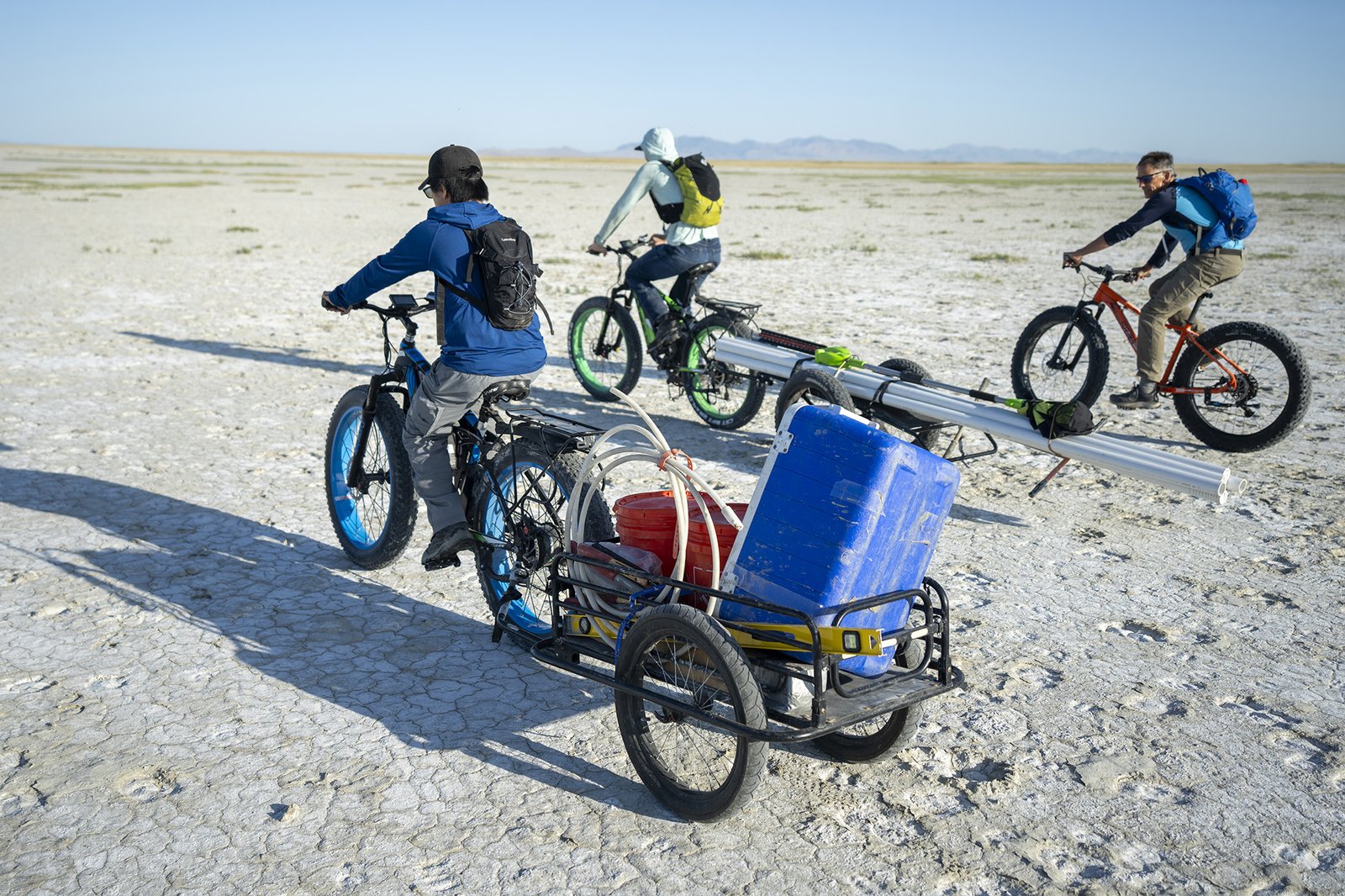 Three cyclists ride across the bed of the Great Salt Lake. Two of the cyclists are hauling research equipment.