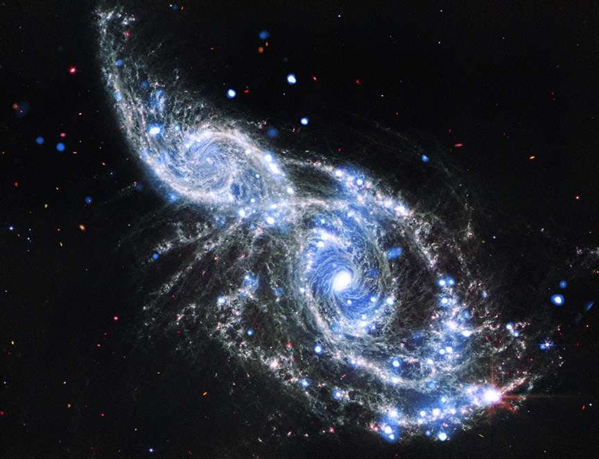 A pair of colliding spiral galaxies. Here, both spirals are shown face on, with the smaller of the two galaxies, IC 2163, at the upper left of the larger galaxy, NGC 2207, which dominates the center and lower right of the image. Both galaxies have long, spiraling, silver blue arms, dotted with specs of blue and red. Toward our upper left, the curving arms overlap, and bend toward their neighbors' core. James Webb mid-infrared image (white, gray, and red) and adds the X-ray view from Chandra (blue).