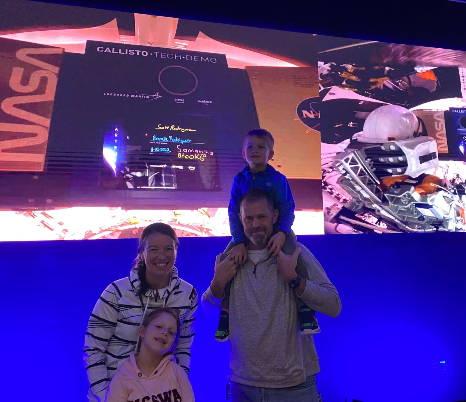 A family of four - mom, dad, and two young children - stand in front of a large screen showing video from inside the Artemis I spacecraft.