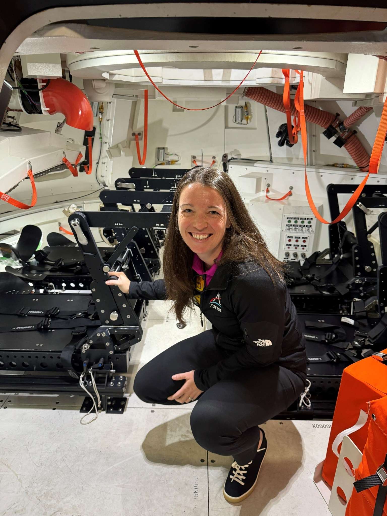 A woman crouches inside the mockup of a spacecraft that is equipped with seats for crewmembers.